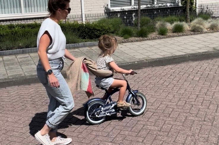 Moeder Simone heeft een sjaal vast en loopt achter haar kind die fietst met een sjaal om zijn middel
